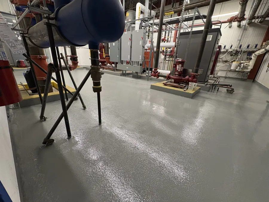 Close view of the broadcast-textured traffic coating in the Canadian Tire mechanical room, terminated around pipe supports and up the face of a plinth