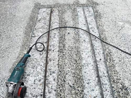 Two saw-cut concrete repairs on a parkade ramp broken out to the rebar, the wire-brush grinder beside them