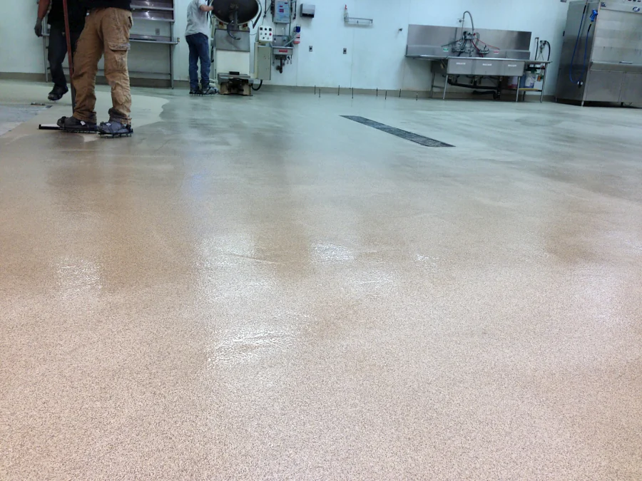 Finished decorative quartz floor across the back-of-house prep room at Costco in Abbotsford, with the sinks, trench drains and stainless equipment beyond