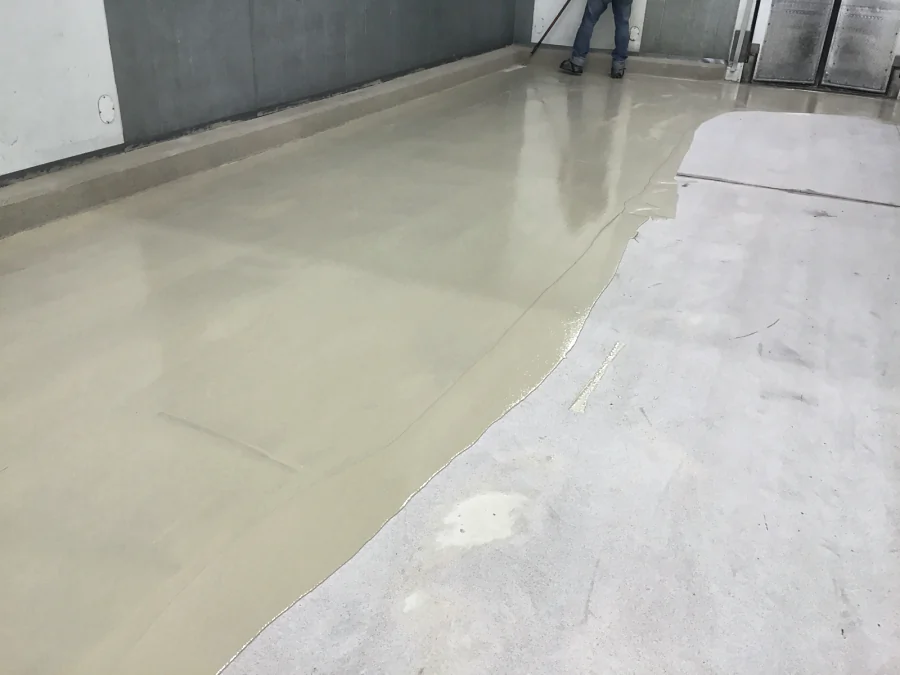 The first tan coat of the decorative quartz floor going down over the prepared slab at Costco Prince George, the coved base already formed along the wall