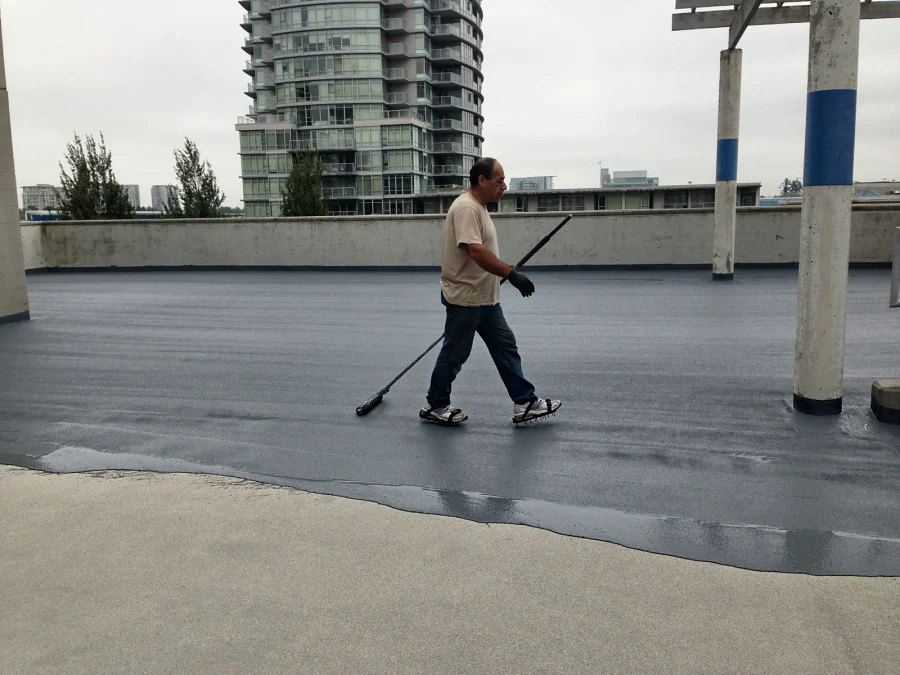 A darker grey coat being rolled out over the lighter coat on the roof deck of the Three West Centre parkade in Richmond
