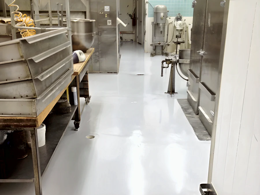 High-gloss light grey floor down the production aisle of a Vancouver bakery, around the round floor drains, with the mixers at the far end